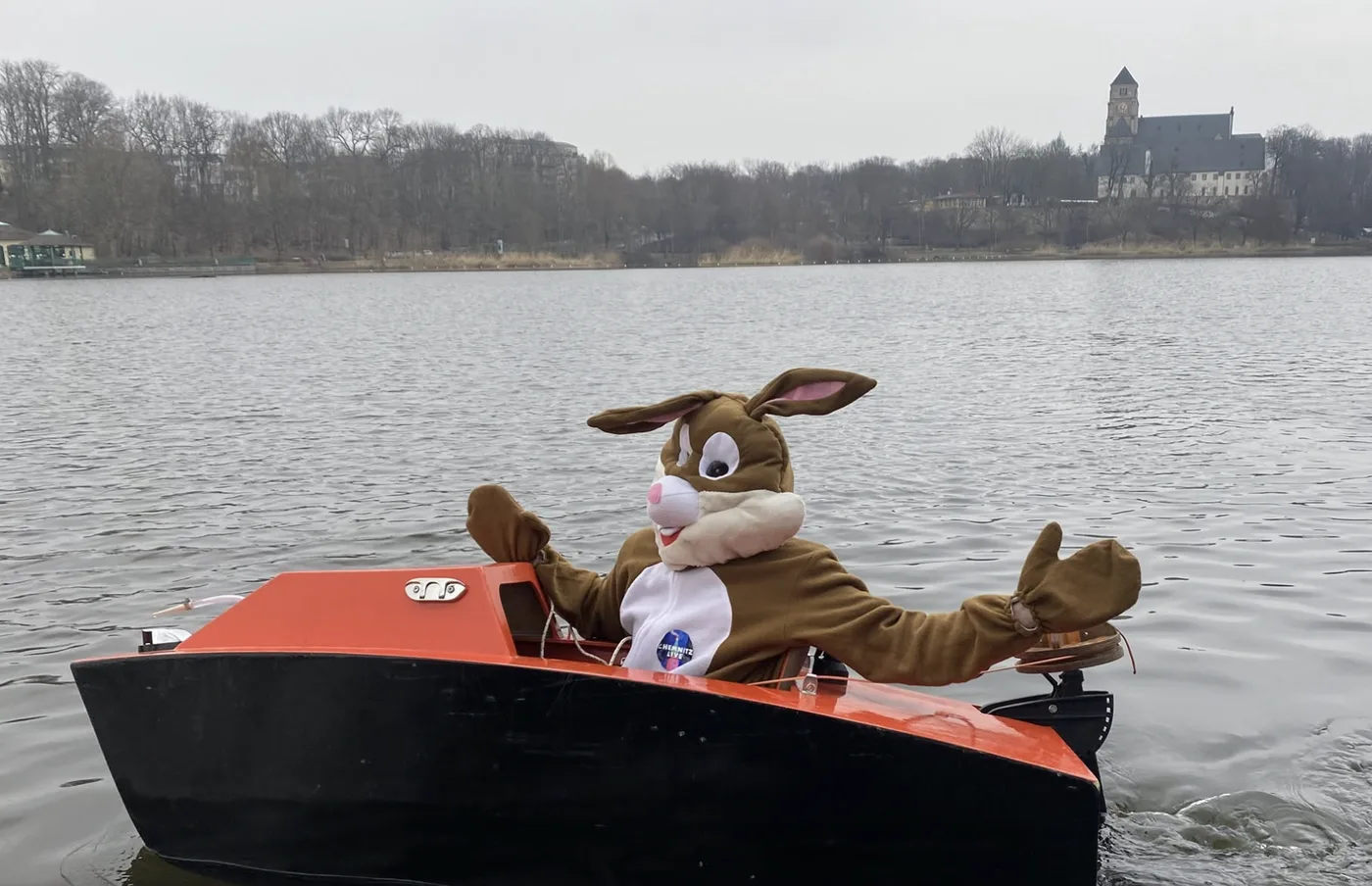 Osterhase im Kostüm winkt aus einem orange-schwarzen Boot auf dem Chemnitzer Schlossteich, im Hintergrund die Schlosskirche