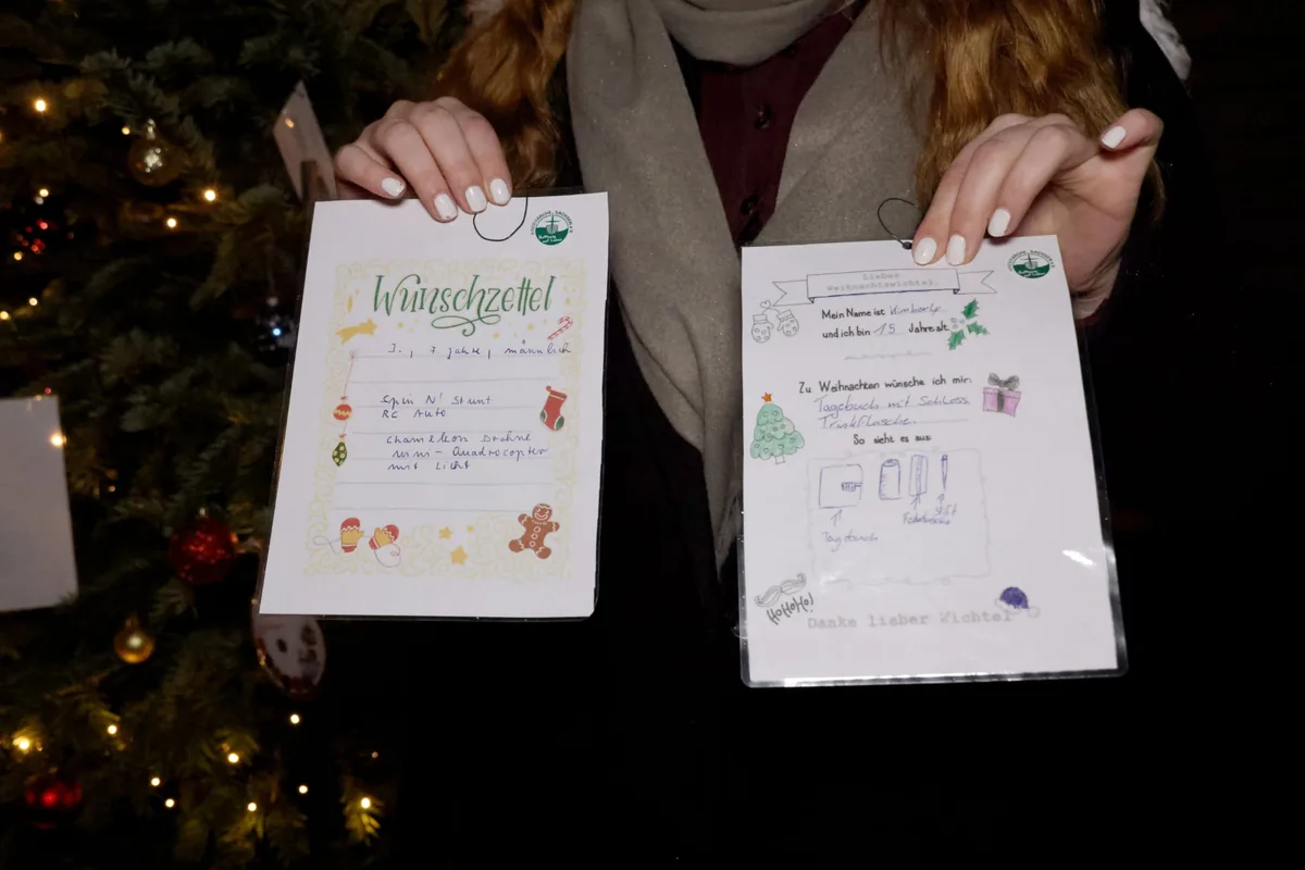 Zwei handgeschriebene Wunschzettel werden vor einem beleuchteten Weihnachtsbaum in die Kamera gehalten