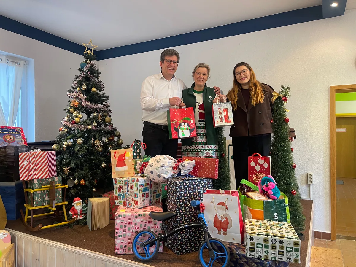 Drei Erwachsene mit Geschenktüten hinter verpackten Geschenken und einem geschmückten Weihnachtsbaum